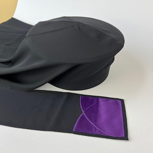 May include: Black head covering with a rounded top and a long, attached scarf. The scarf has a rectangular purple accent with a decorative design. The head covering is made of a smooth, dark fabric.