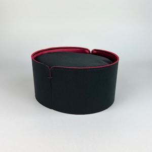 May include: A black hat with a cylindrical shape and a red interior lining. The hat has a rounded top and a small slit on the side. The hat is made of a smooth, matte material.