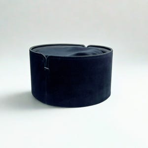 Może przedstawiać: Cylindryczny, czarny aksamitny kapelusz z płaskim wierzchem i ciemnym wnętrzem. Kapelusz ma strukturalny kształt i subtelny połysk, na tle gładkiego, białego tła. Design kapelusza sugeruje formalny lub tradycyjny styl.