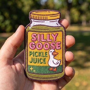 May include: An embroidered patch shaped like a jar with a yellow label featuring the text SILLY GOOSE PICKLE JUICE in pink and green letters. The design includes a white goose with an orange beak and feet, plus small yellow star accents.