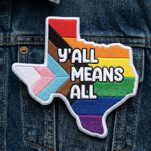 May include: Embroidered patch in the shape of Texas, featuring a rainbow design and the text "Y'all Means All." The patch includes the transgender flag colors and a black outline. It is set against a denim jacket.