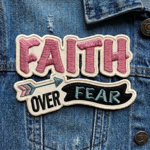 May include: Embroidered patch with the words "FAITH OVER FEAR". The word "FAITH" is in pink with a white border. "OVER" is on an arrow, and "FEAR" is on a black banner, both in light blue.