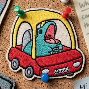 May include: Embroidered patch featuring a teal creature with an open mouth, inside a red car with a yellow top. The patch has the word "AHHH!" on the front. The patch is attached to a corkboard with push pins.