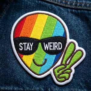May include: Embroidered patch featuring a rainbow-colored alien with sunglasses and the words "STAY WEIRD." The alien has a smiling face and a peace sign hand gesture. The patch is on a denim background.