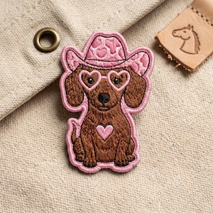 May include: Embroidered patch featuring a brown dachshund wearing a pink cowboy hat with heart patterns and heart-shaped glasses. A pink heart is on the dog's chest. The patch has a pink border and is on a beige background.