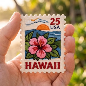May include: A rectangular embroidered patch shaped like a postage stamp featuring a pink hibiscus flower, green leaves, a blue ocean, and an orange sun. The text 25 USA and HAWAII is stitched in red. The patch has a cream-colored border with scalloped edges.