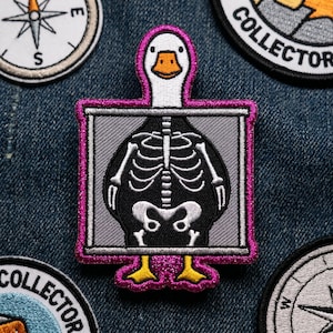 May include: Embroidered patch featuring a white duck with an orange beak and feet, framed in a pink border. The duck's body is a black x-ray showing its skeleton. Other patches with compass and collector text are visible.