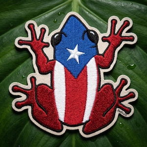 May include: Embroidered patch featuring a frog design with the flag of Puerto Rico. The frog has a blue head with a white star, red and white stripes on its body, and red limbs. The patch is outlined in beige.