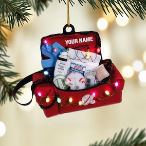 May include: A red Christmas ornament shaped like a couch, filled with medical supplies and a blue medical device. The ornament has a black handle and is decorated with colorful lights. The text "YOUR NAME" is printed on the top.