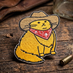 May include: Embroidered patch featuring a yellow cat wearing a brown cowboy hat and a red bandana with a white floral pattern. The cat is sitting with its legs tucked under. The patch has a black border.
