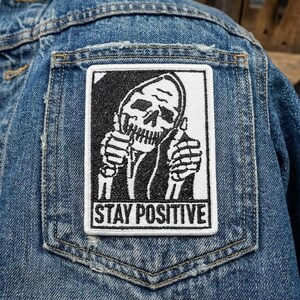 May include: A denim jacket with a rectangular patch featuring a black and white illustration of a skull giving a thumbs-up. The text "STAY POSITIVE" is below the skull.