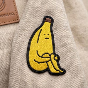 May include: A yellow banana-shaped embroidered patch with a smiling face and crossed legs. The patch has a black outline and brown top, sewn onto a beige fabric. A brown leather tag with text is visible in the background.