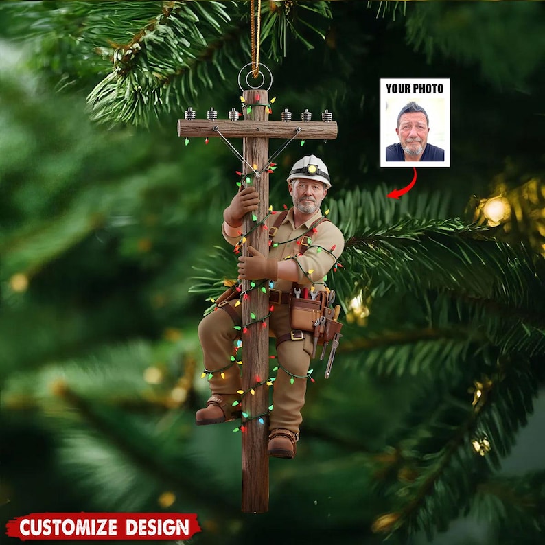 Puede incluir: Adorno navide&ntilde;o que representa a un liniero subiendo a un poste de electricidad decorado con luces de colores. El liniero lleva casco, ropa de trabajo y cintur&oacute;n de herramientas. El adorno tiene una cuerda dorada para colgar y un espacio para una foto. El texto "CUSTOMIZE DESIGN" est&aacute; en la parte inferior.