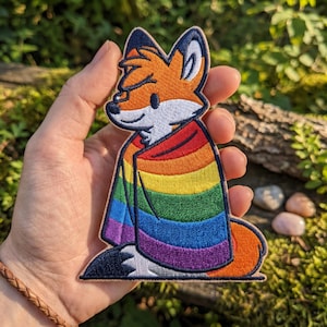 May include: Embroidered patch featuring a cartoon fox with orange fur, white face, and a rainbow-colored cape. The patch has a black border and is held against a natural, green background.