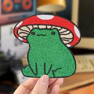 May include: Embroidered patch featuring a green frog under a red mushroom cap with white spots. The frog has a smiling expression and is outlined in black. The patch is likely for clothing or accessories.