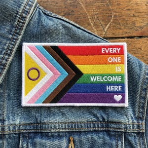 May include: Embroidered patch with a rainbow flag design, featuring the words "EVERY ONE IS WELCOME HERE" and a white heart. The patch includes the intersex flag and the transgender flag. The patch is on a denim jacket.