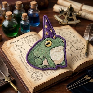 May include: Embroidered patch featuring a green frog wearing a purple wizard hat with gold stars. The patch is set on an open book with the text "SPILLS OF THE CROAKING ONE". The background includes potion bottles and scrolls.