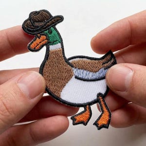 May include: Embroidered patch of a duck wearing a brown cowboy hat. The duck has a green head, brown body, white chest, and orange feet and beak. The patch is held between two hands.