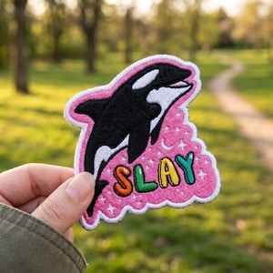 May include: Embroidered patch featuring a black and white orca whale design on a pink background. The word "SLAY" is written in colorful letters. The patch has a white border and is decorated with stars and a crescent moon.