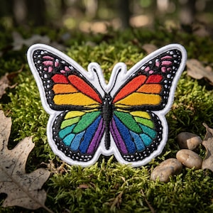 May include: Embroidered butterfly patch with a rainbow design. The butterfly has a white border and black detailing on the wings. The wings are filled with vibrant colors, including red, orange, yellow, green, blue, and purple. The patch is set against a background of green moss.