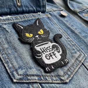 May include: Black cat patch with yellow eyes and the text "Hiss Off" on a white mug. The patch is embroidered and sewn onto a denim jacket pocket. The cat is holding the mug with both paws.