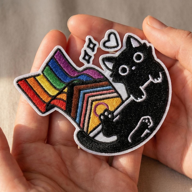 May include: Embroidered patch featuring a black cat playfully interacting with a rainbow flag and a progress pride flag. The patch has a white border and includes heart and star-shaped details. The cat has large, round eyes.