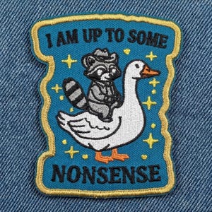 May include: Embroidered patch with a teal background and gold trim. The patch features a raccoon riding a white goose, with the text "I AM UP TO SOME" above and "NONSENSE" below. Gold stars are scattered around the image.