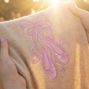 Puede incluir: Un suéter beige con un diseño bordado en rosa de zapatillas de ballet y un lazo. Las zapatillas de ballet están detalladas con cintas y son de color rosa suave. El suéter se sostiene frente a un fondo brillante y soleado.