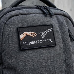 May include: A dark gray backpack with a black patch featuring an embroidered design of two hands, one human and one skeletal, with the words "MEMENTO MORI" below. The backpack has a top handle and a zipper.