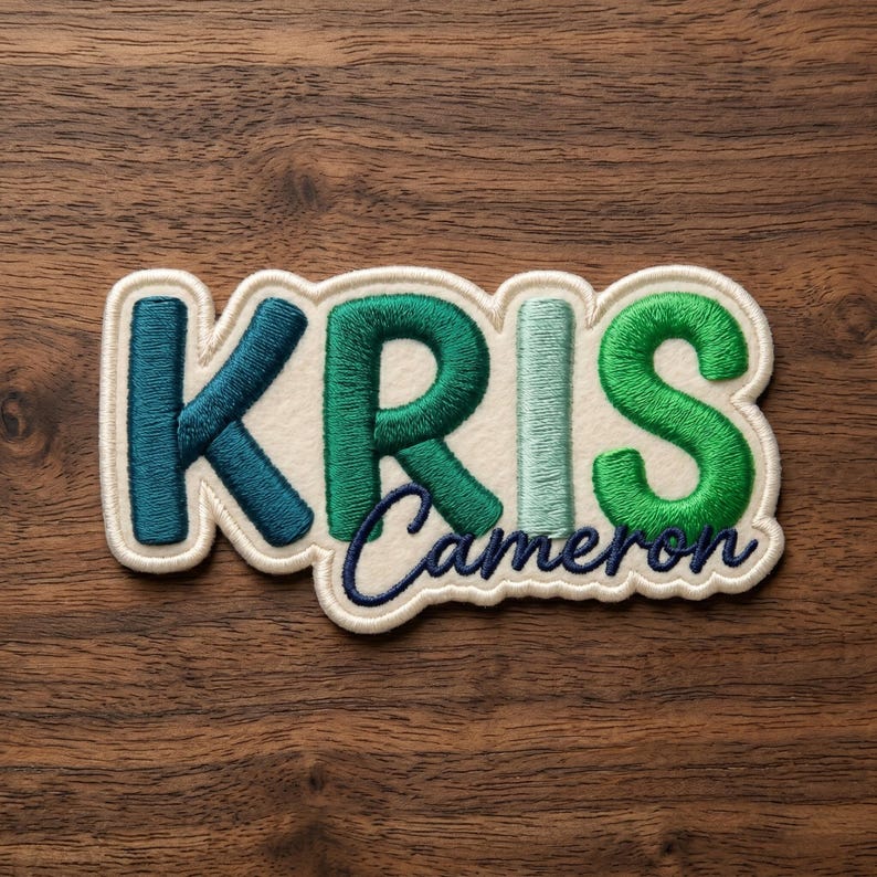 May include: A custom embroidered patch featuring the name KRIS in large, block-style letters with a gradient of teal, dark green, and light green thread, with the name Cameron embroidered in dark blue cursive script underneath on a cream felt base.