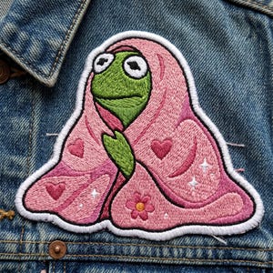 May include: Embroidered patch featuring a green frog character wrapped in a pink blanket. The blanket is decorated with hearts, stars, and a flower. The patch has a white border and is set against a denim jacket.
