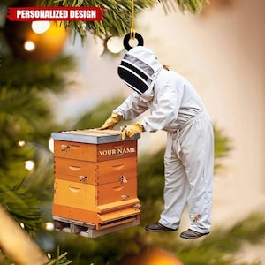 May include: A personalized ornament featuring a beekeeper tending to a wooden beehive. The ornament has the text "PERSONALIZED DESIGN" at the top and "YOUR NAME" on the hive. The beekeeper is wearing a white suit and gloves. The beehive is orange and yellow.