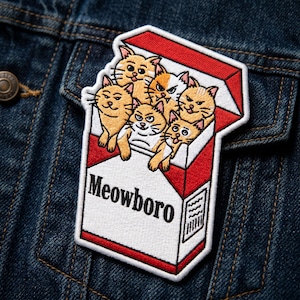 May include: Embroidered patch featuring a cigarette pack design with the text "Meowboro." The pack is red and white, with several cartoon cats peeking out. The patch is attached to a denim jacket.