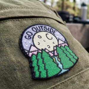 May include: Embroidered patch with the words "GO OUTSIDE" in a circular design. The patch features a smiling moon, mountains, and green trees. The patch is attached to a green fabric.