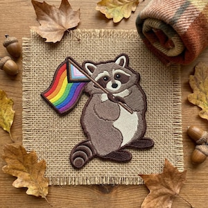 May include: Embroidered patch featuring a raccoon holding a rainbow flag with a transgender pride stripe. The raccoon is brown and white, with a black mask and a neutral expression. The patch is on a burlap surface, surrounded by fall leaves and acorns.