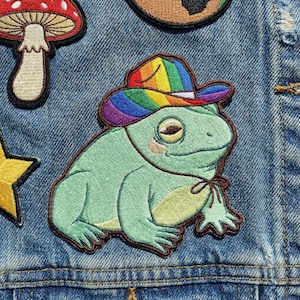 May include: Embroidered patch featuring a light green frog wearing a rainbow cowboy hat. The frog has a light brown outline and a small bow tie. Other patches, including a mushroom and star, are visible on a denim background.