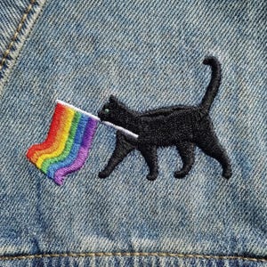 May include: Embroidered patch featuring a black cat carrying a rainbow flag. The cat has green eyes and is stitched onto a blue denim fabric. The rainbow flag has stripes of red, orange, yellow, green, blue, and purple.