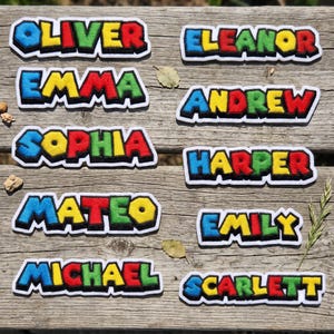 May include: A collection of nine embroidered name patches featuring the names Oliver, Eleanor, Emma, Andrew, Sophia, Harper, Mateo, Emily, Michael, and Scarlett. Each letter is rendered in a bold, multi-colored block style with a black outline.
