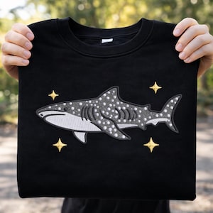 Puede incluir: Sudadera negra con un diseño bordado de tiburón ballena con puntos blancos y detalles grises. El diseño incluye tres acentos de estrellas doradas. La sudadera es sostenida por una persona.