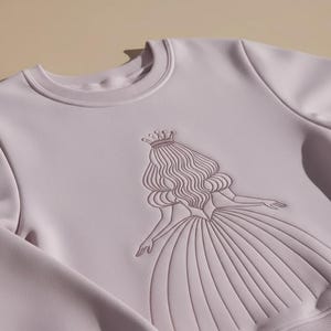 May include: A light purple sweatshirt featuring an embroidered design of a princess wearing a crown and gown. The princess design is in a darker shade of purple. The sweatshirt has a crew neck and long sleeves.