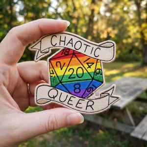 May include: Embroidered patch featuring a rainbow-colored D20 die with the words "CHAOTIC" and "QUEER" on banners. The die has numbers in yellow and green. The patch is held up against a blurred outdoor background.