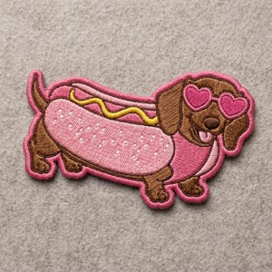 May include: Embroidered patch featuring a dachshund dog in a hot dog bun. The dog is brown with pink heart-shaped sunglasses and a pink outfit. The hot dog bun is pink with white seeds and yellow mustard.