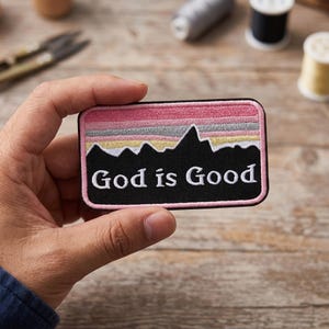 May include: Embroidered patch with a black mountain range silhouette and the text "God is Good" in white. The patch features a pink and gray sunset design above the mountains, with a black border.