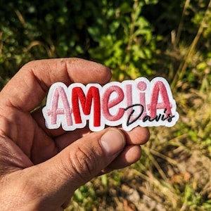 May include: A custom embroidered patch featuring the name Amelia in a mix of light pink, red, and dark pink thread, with the surname Davis stitched in black cursive script below the letters i and a, all set against a white fabric background.