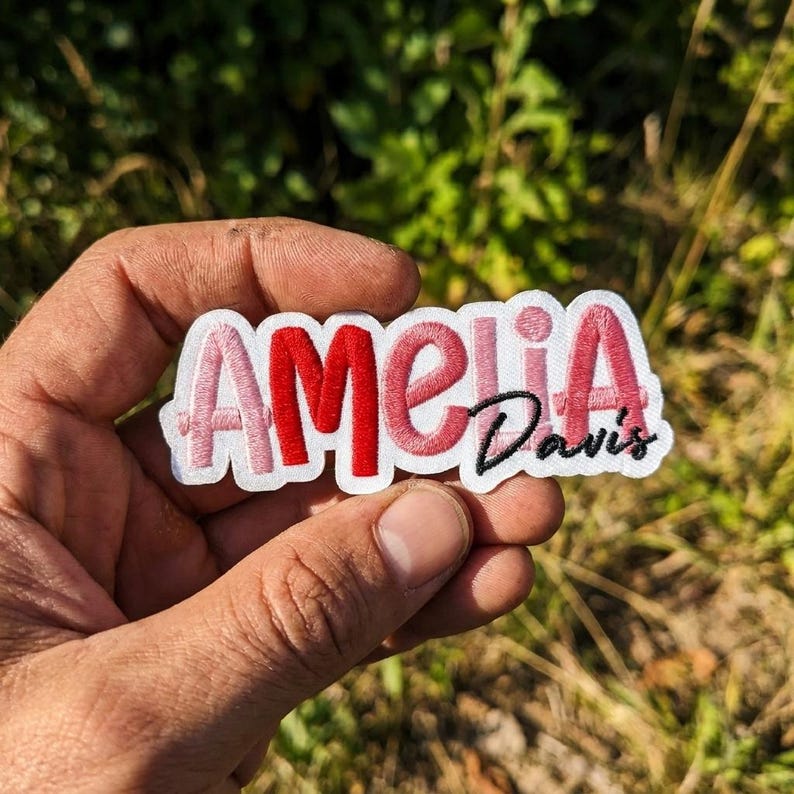 May include: A custom embroidered patch featuring the name Amelia in a mix of light pink, red, and dark pink thread, with the surname Davis stitched in black cursive script below the letters i and a, all set against a white fabric background.