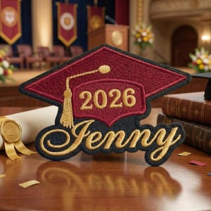 May include: A maroon and black graduation cap patch with the name "Jenny" embroidered in gold, along with the year "2026". The patch is on a wooden surface, with a diploma and books in the background.
