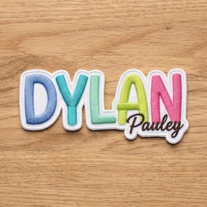May include: A custom embroidered patch featuring the name DYLAN in large, multi-coloured block letters including blue, teal, mint green, lime green, and pink, with the name Pauley embroidered in smaller brown script lettering below.