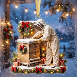 May include: A clear acrylic Christmas ornament featuring a beekeeper in a white protective suit tending to a wooden beehive. The hive is decorated with two small green wreaths with red bows, the name Wilson, and wrapped gift boxes at the base.