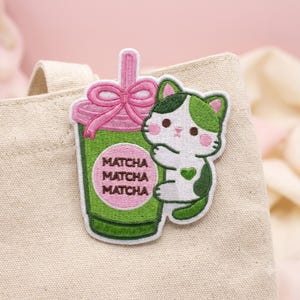 May include: An embroidered patch featuring a green matcha drink in a cup with a pink bow and straw, alongside a white and green cat with a small green heart on its chest. The cup has a pink circular label with the text MATCHA MATCHA MATCHA.