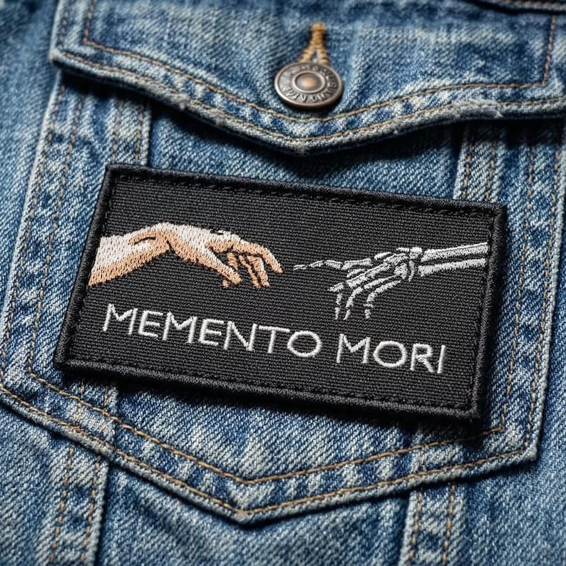 May include: Black rectangular patch with the words "MEMENTO MORI" in white, featuring a human hand and a skeletal hand reaching towards each other. The patch is sewn onto a blue denim jacket.
